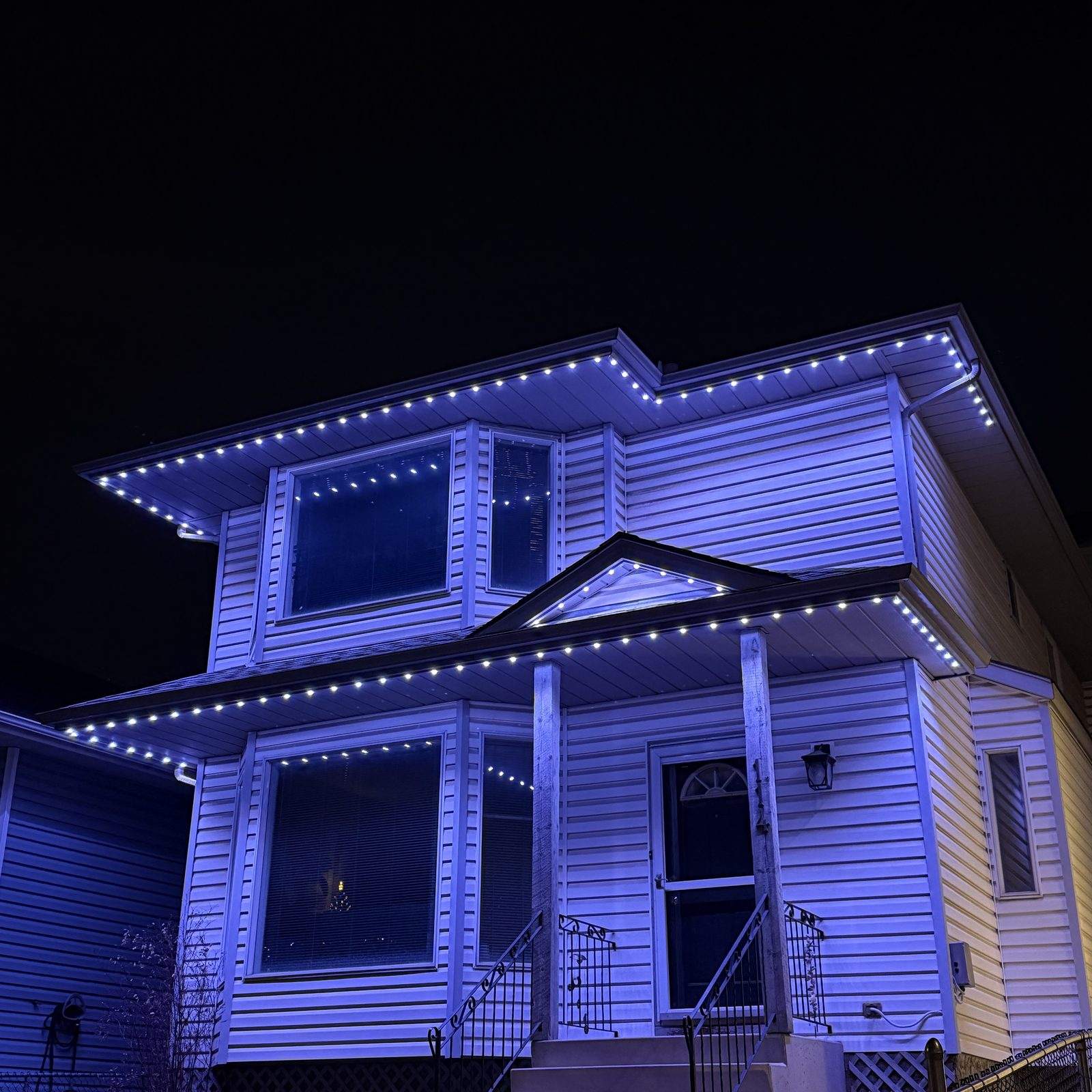 Permanent Christmas lights running blue along the roofline, covered porch and window trim of a white two-storey home at night
