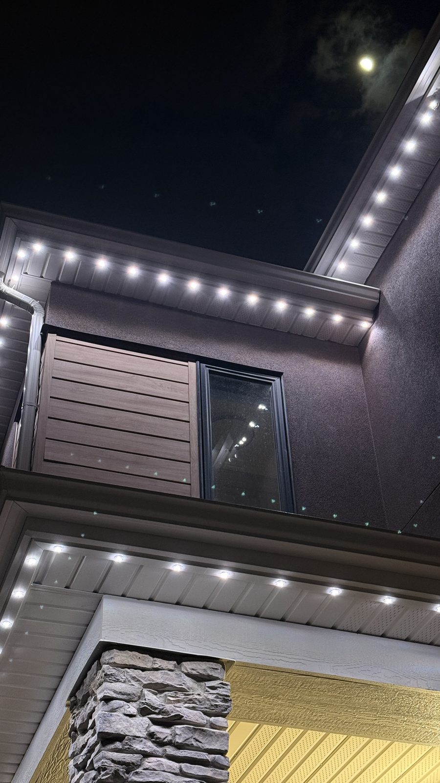 Cool white permanent lighting along the stacked soffits of a grey stucco and stone home under a moonlit sky