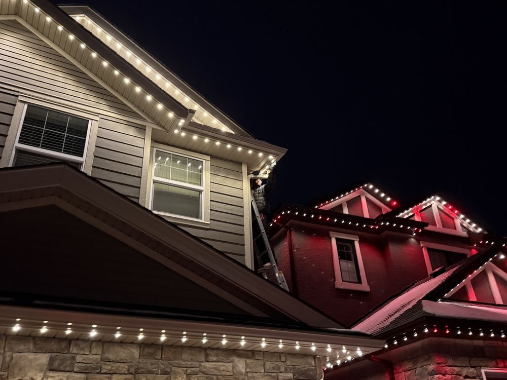 A Luminair installer on a ladder fitting track to a fascia board at night, with finished rooflines lit behind