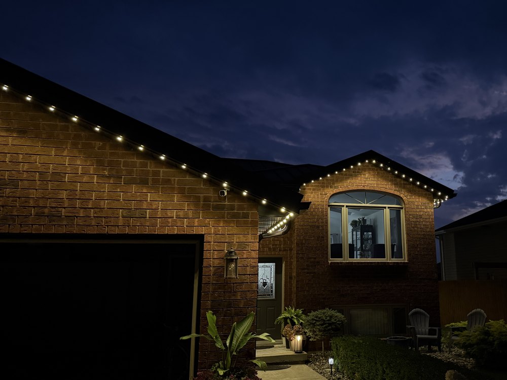Warm white permanent lighting following the garage gable and front gable of a brick home against a deep blue dusk sky