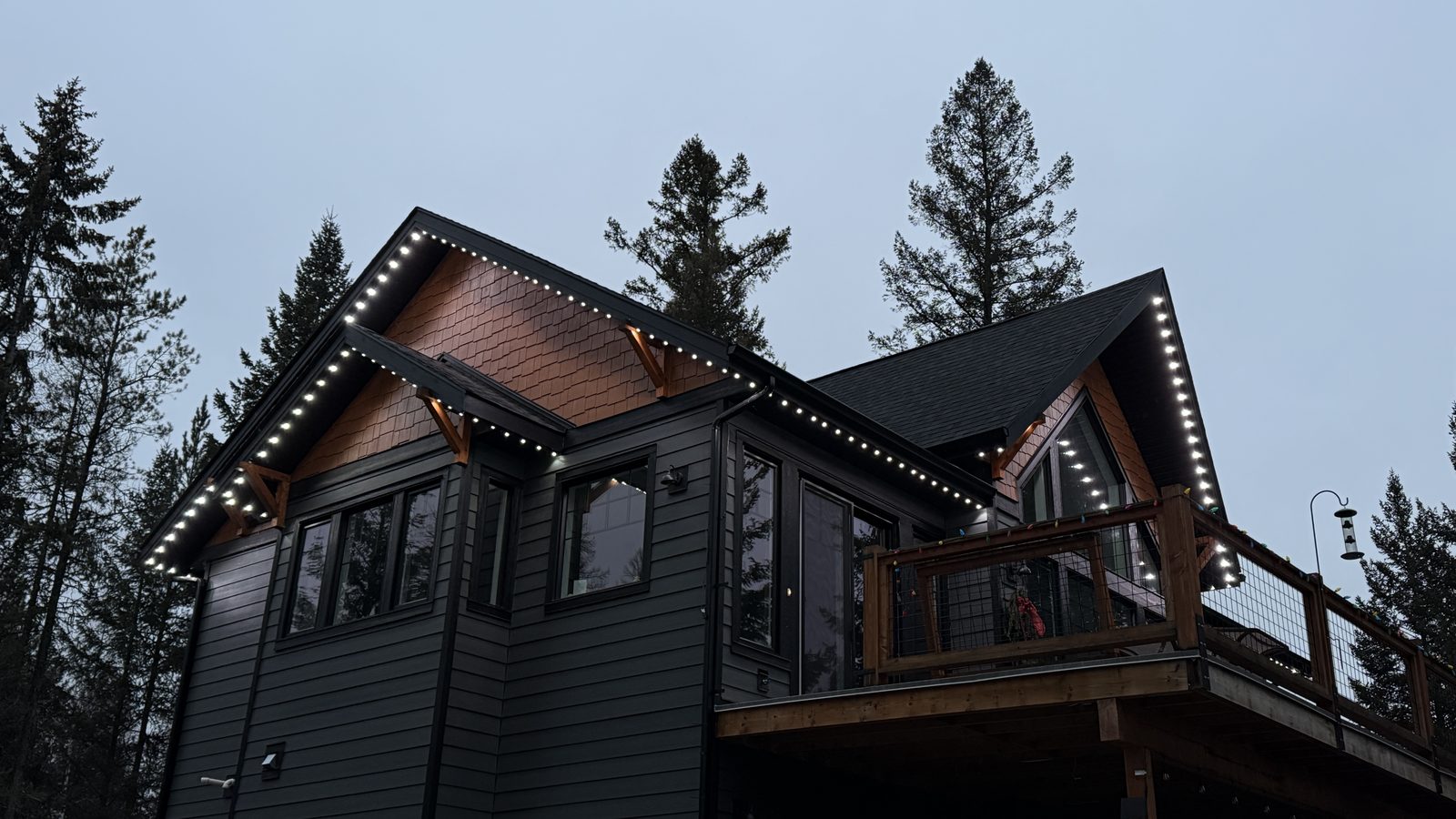 Warm white permanent lighting following the steep gables of a dark cedar-shingled chalet surrounded by evergreens at dusk