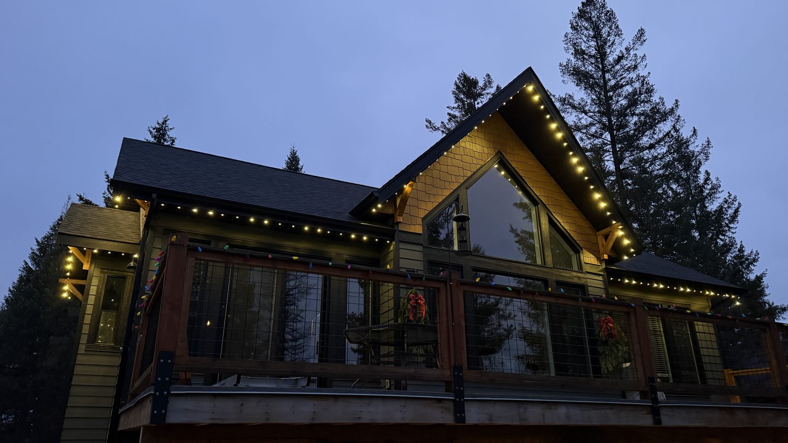 The full front elevation of a timber-framed chalet outlined in warm white above a wraparound deck at dusk