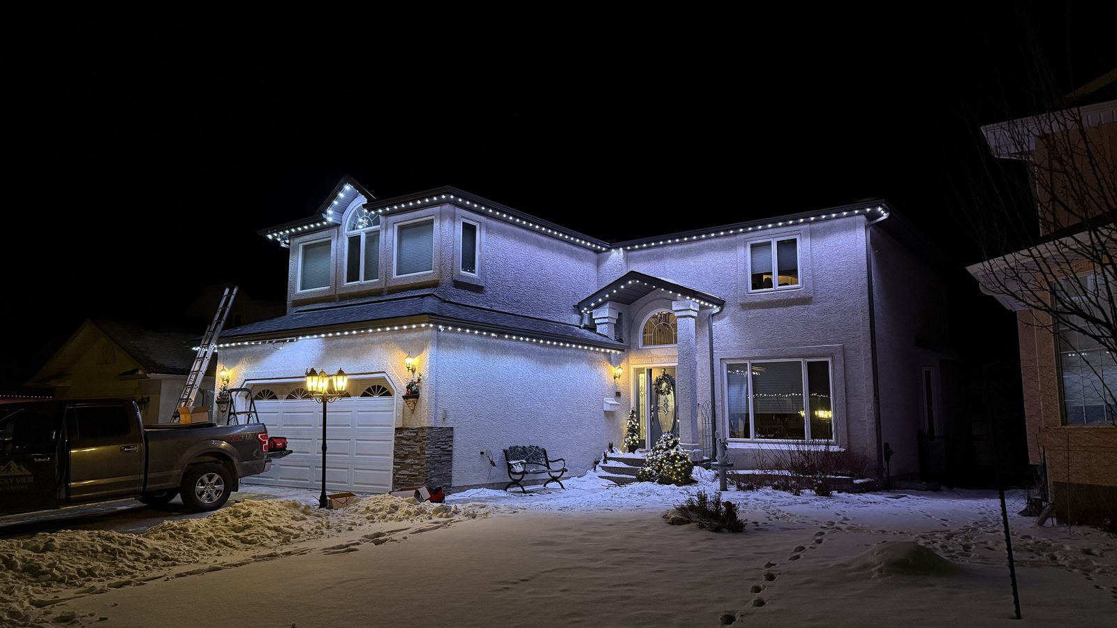 A completed warm white installation on a stucco two-storey at night, with the installation truck and ladder still on the driveway